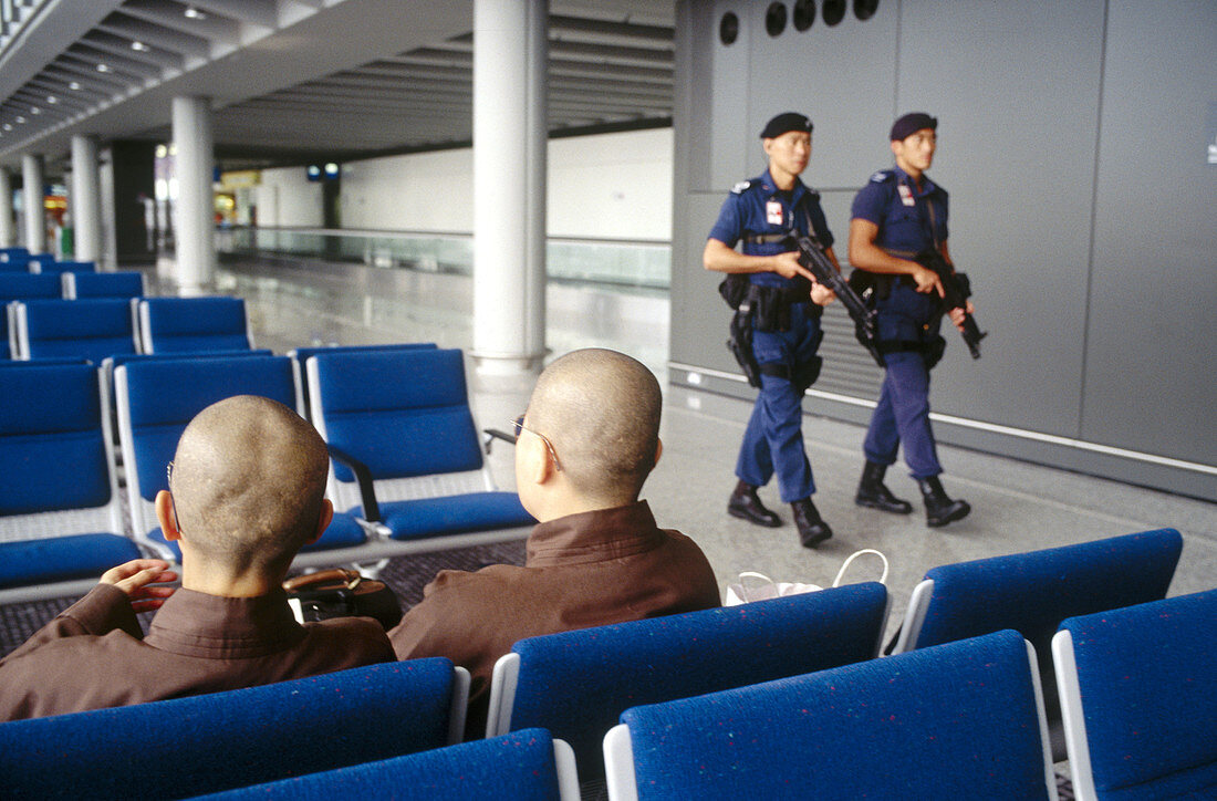 Two armed guards with assault machineguns patrol the Hong Kong International Airport, passing two female Buddhist nuns.