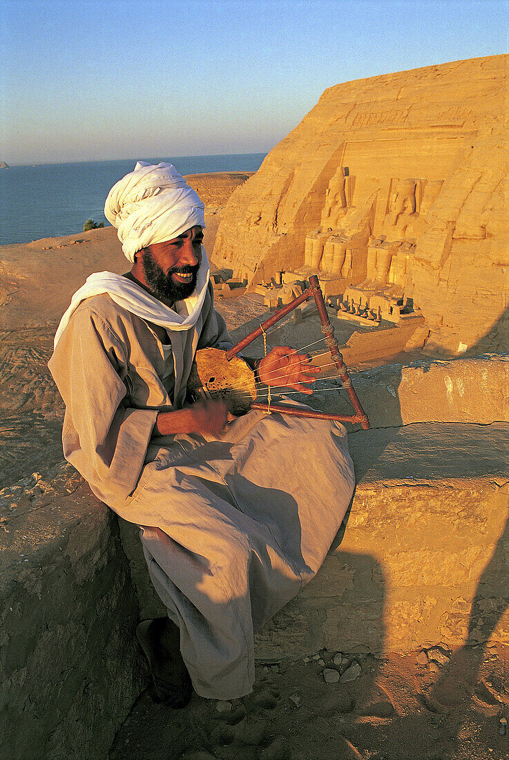 Bearded man playing sithar at the sunrise, Abou Simbel Ramasses temple at back. Nubia . Egypt.