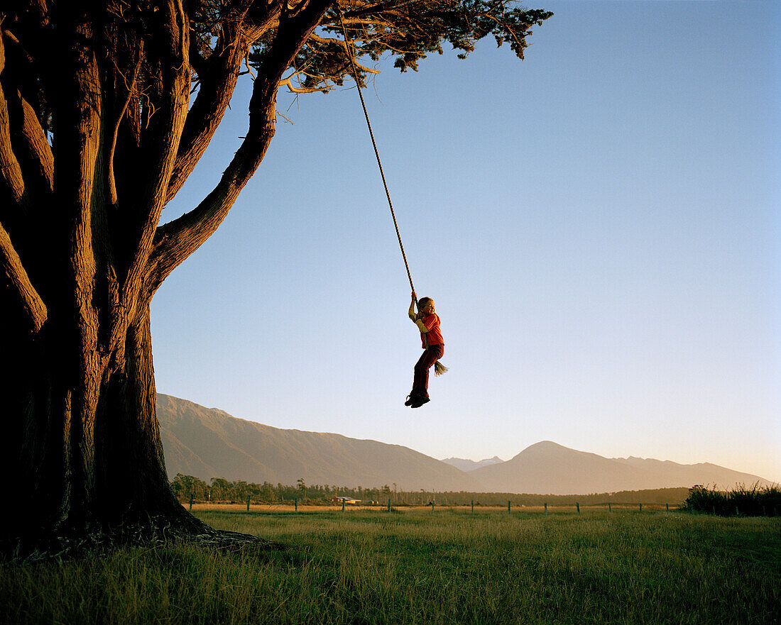 Mädchen auf Schaukelseil an riesigem Baum bei Sonnenuntergang, Westküste, Südinsel, Neuseeland