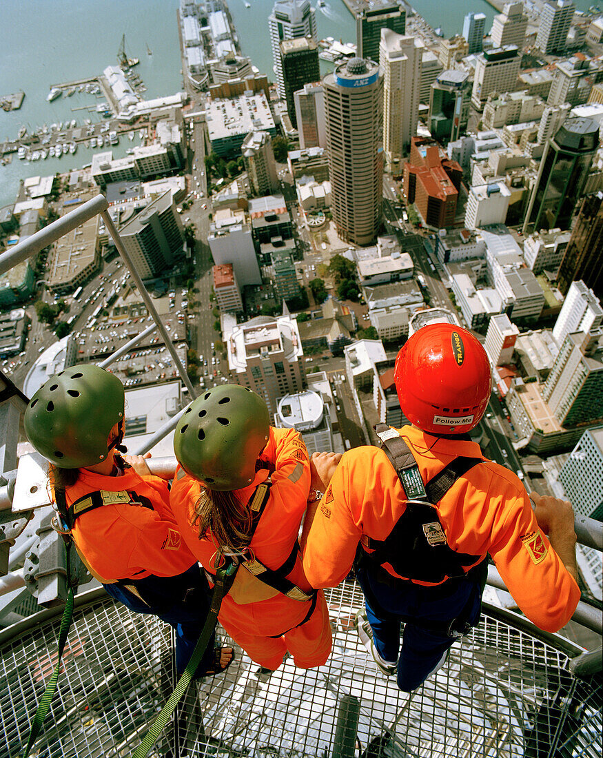 Drei Personen mit Kletterausrüstung auf dem Sky Tower schauen über den Central Business District und Waitemata Harbour, Auckland, Nordinsel, Neuseeland