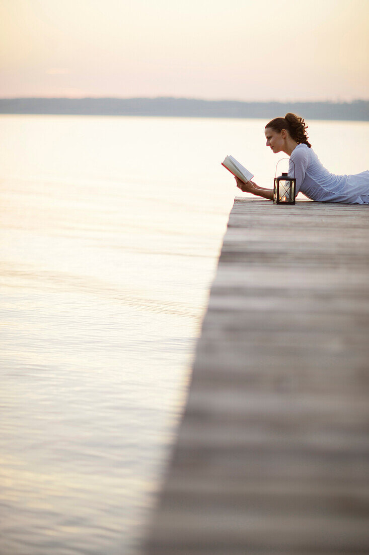 Frau liegt auf einem Steg am Starnberger See und liest ein Buch, Ambach, Bayern, Deutschland