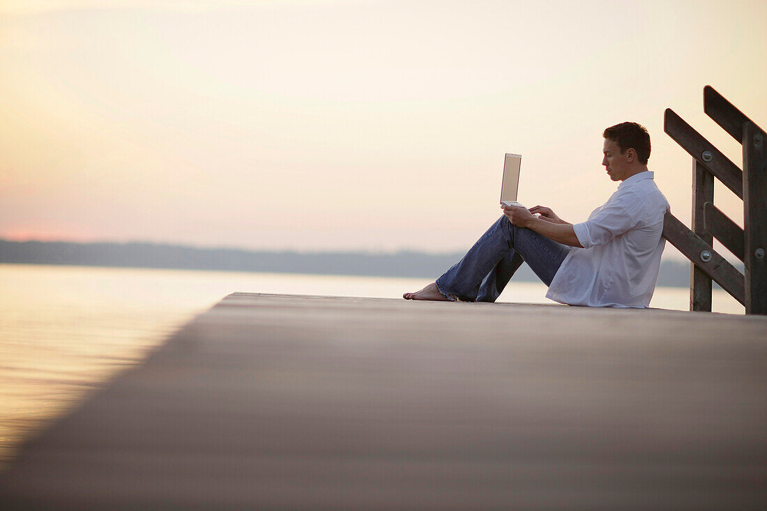 Man using a laptop while sitting on a jetty at lake Starnberg, Ambach, Bavaria, Germany