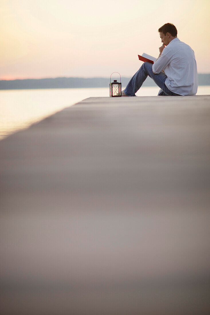 Mann sitzt auf einem Steg am Starnberger See und liest, Ambach, Bayern, Deutschland