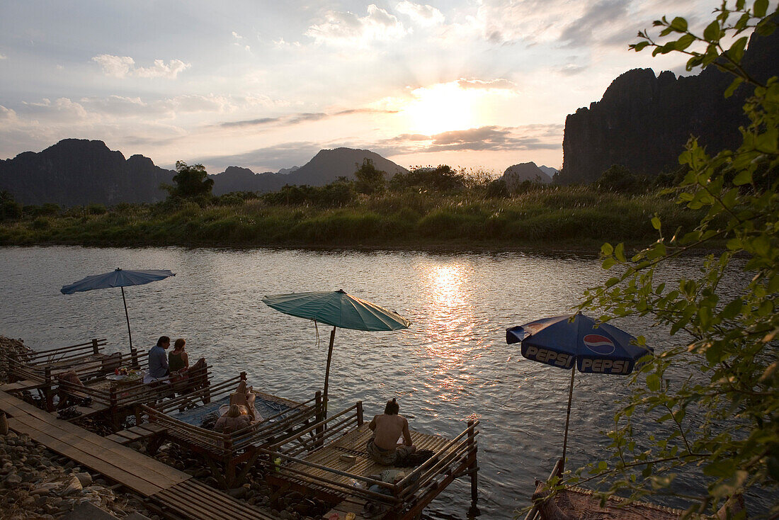 Menschen entspannen sich auf Holzstegen am Fluss Nam Xong bei Sonnenuntergang, Vang Vieng, Provinz Vientiane, Laos