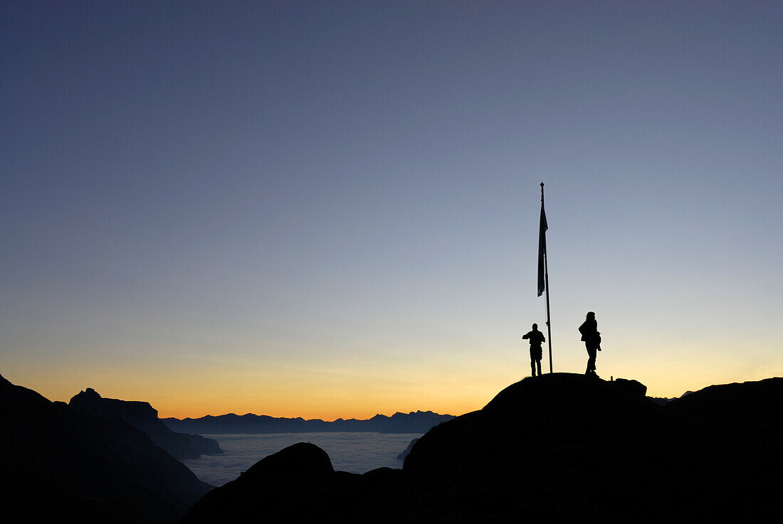 two hikers on dome with flagpole, dawn above fog bank in valley Gschnitztal, Kirchdachspitze in background, Bremer Huette, Stubaier Alpen range, Stubai, Tyrol, Austria