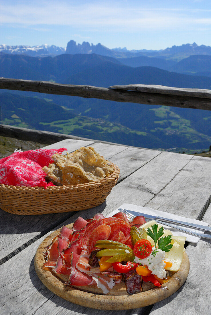 South Tyrolean snack with South Tyrolean speck, bacon, salami, peperoni, pepperoni, Kaminwurzen, cheese and Schuettelbrot, Dolomites in background, terrace of hut Radlseehuette, Sarntaler Alpen, Sarntal range, South Tyrol, Italy