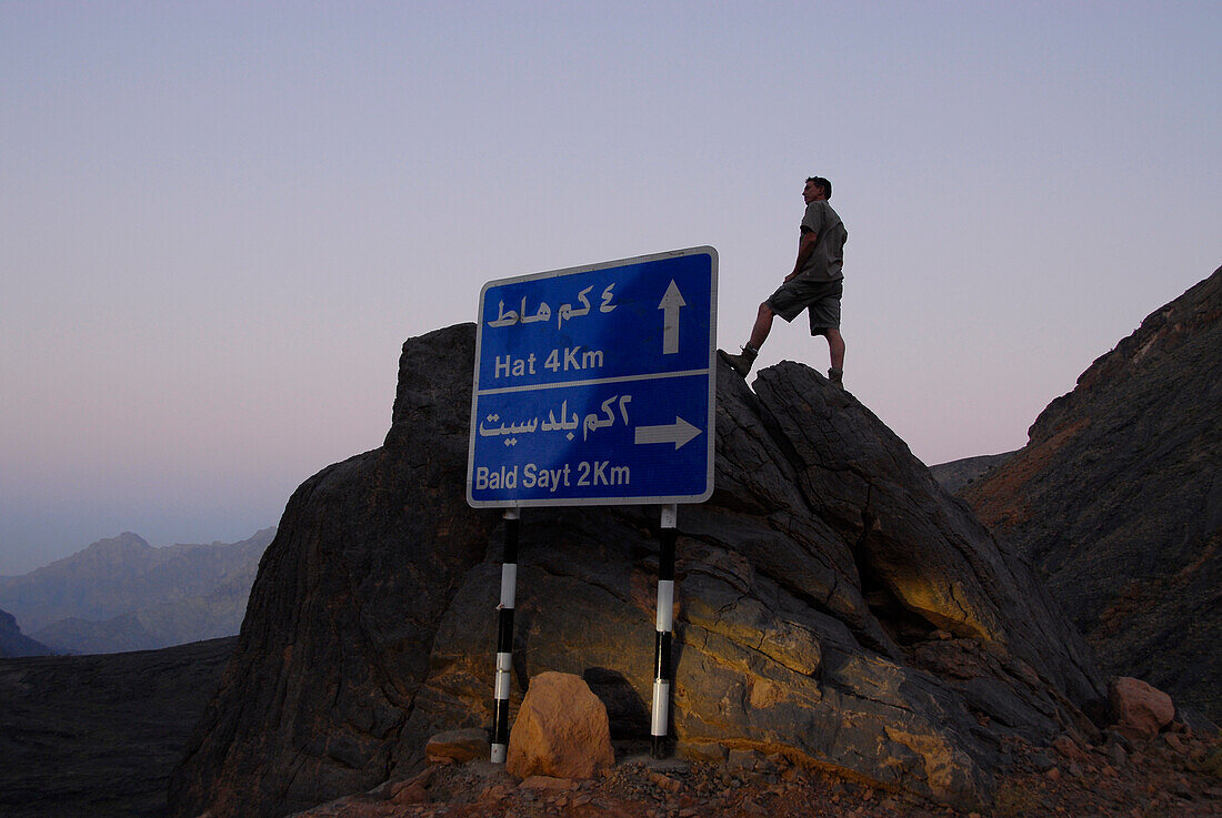 A road sign and man on a rock at dawn, Al Hajar mountains, Oman, Asia