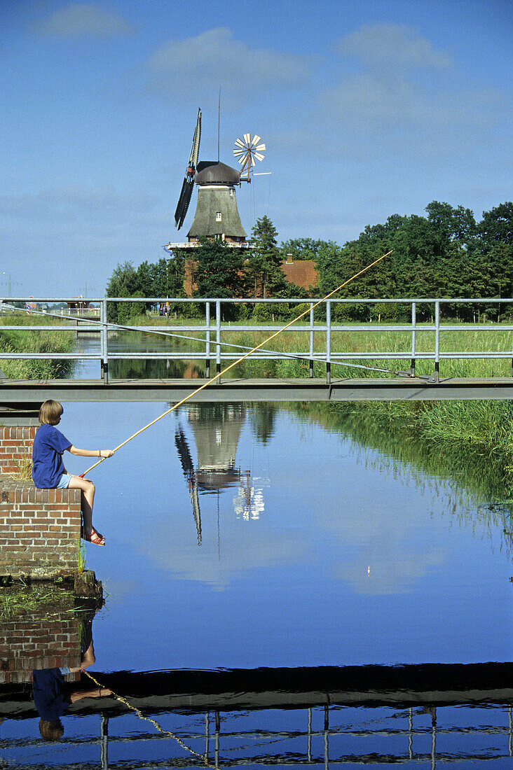 Kind angelt an einem Kanal, Windmühle im Hintergrund, Rhauderfehn, Niedersachsen, Deutschland