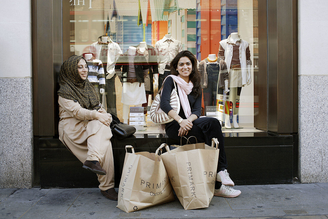 Two beautiful and young moslem ladies sitting in front of the window of a department store, taking a break from a long shopping trip and having their shopping bags in front of them, Oxford Street, London, United Kingdom, Europe