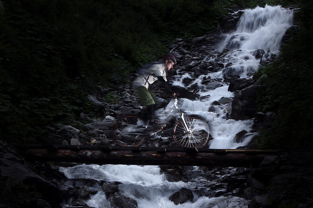 Mountainbiker fährt vor einem Wasserfall über eine Brücke, Ischgl, Tirol, Österreich