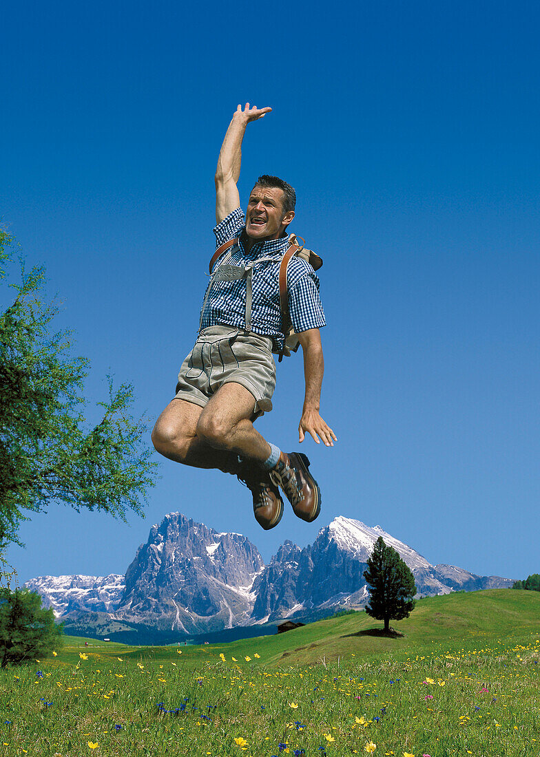 Ein Mann mit Lederhosen macht einen Luftsprung auf einer Almwiese, Seiser Alm, Südtirol, Italien, Europa
