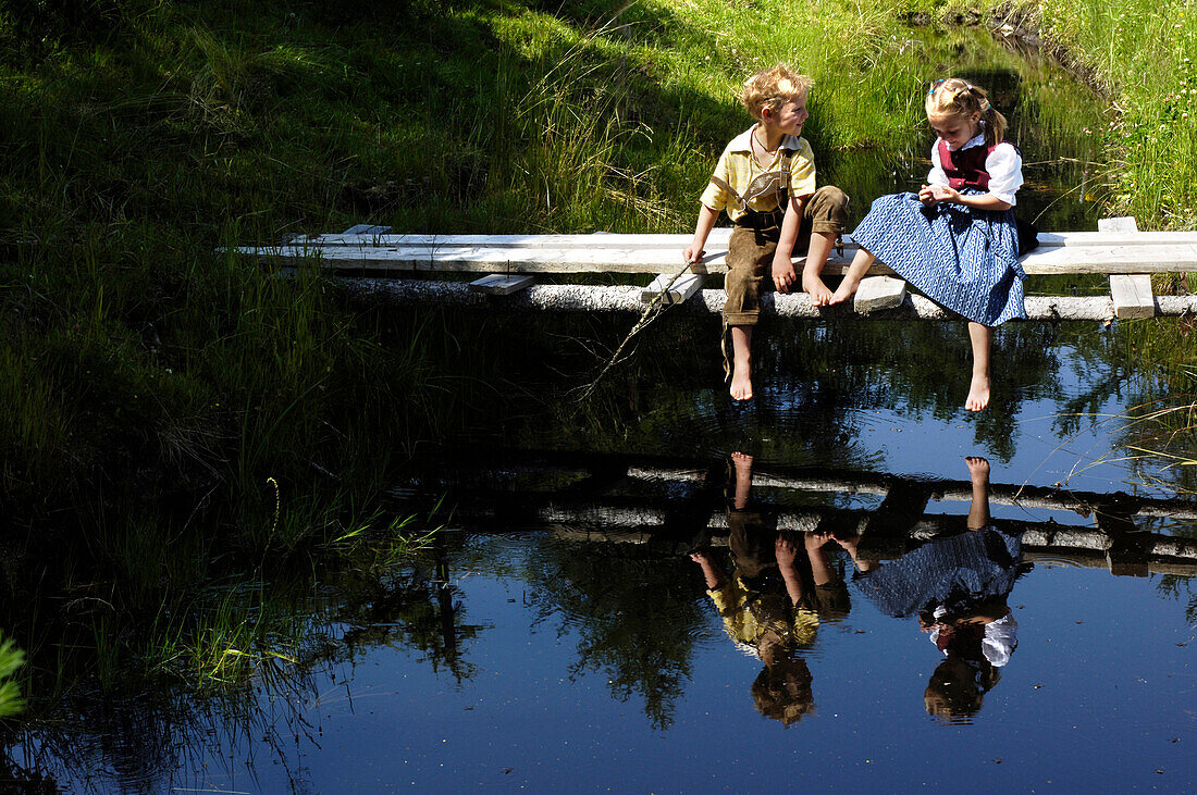 Boy and girl in traditional dress sitting on a wooden jetty at a lake, Alp, South Tyrol, Italy