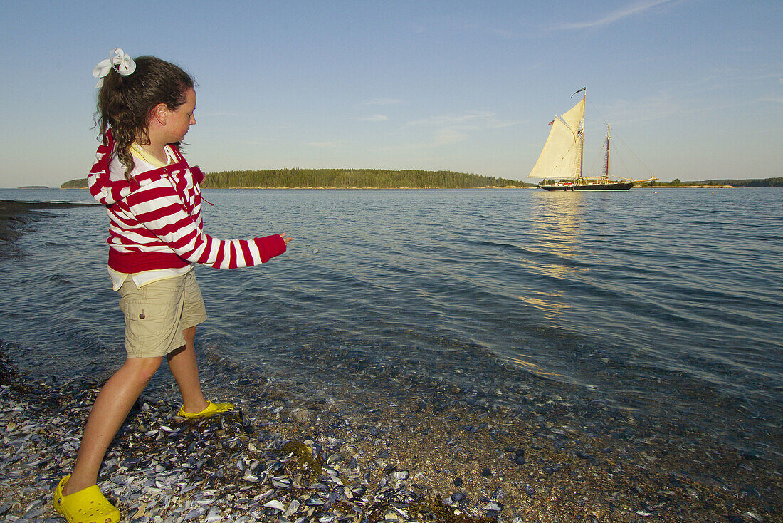 Girl skipping stones across the water with the Schooner Nathaniel Bowditch moored behind, Russ Island, Penobscot Bay, Maine USA