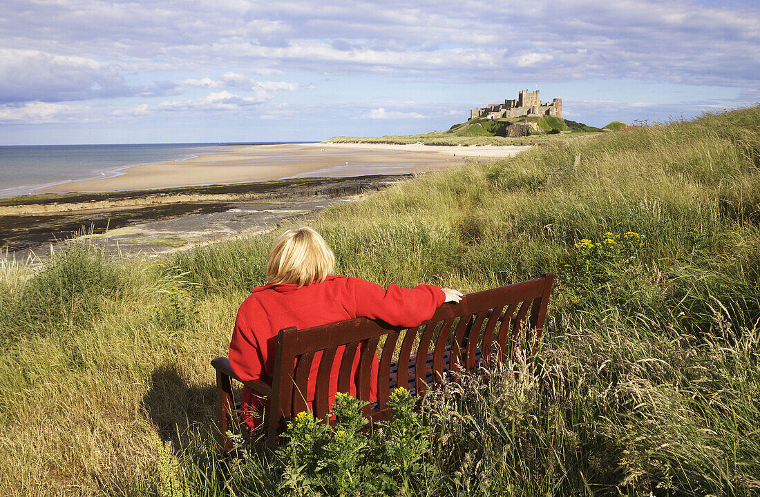 Bamburgh Beach and Castle, with woman wearing a red jacket seated in the foreground, Northumberland, England, UK