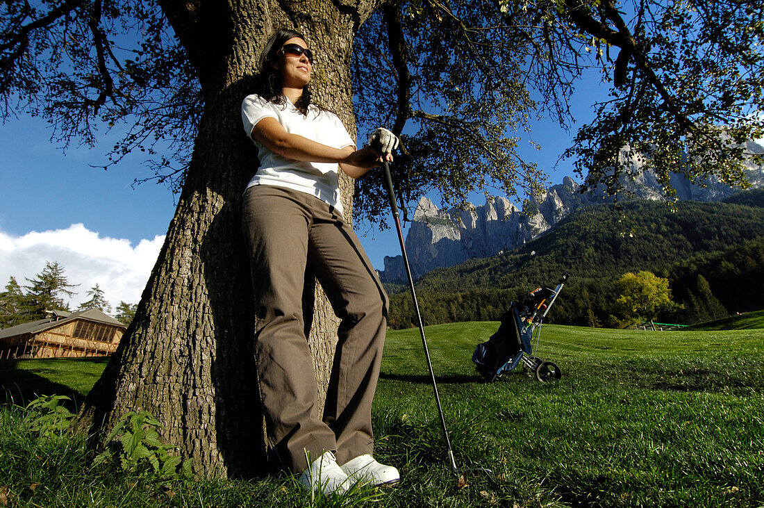 A woman with golf club and sunglasses leaning on a tree, golf court Kastelruth Alpe di Siusi, Sciliar, South Tyrol, Italy, Europe