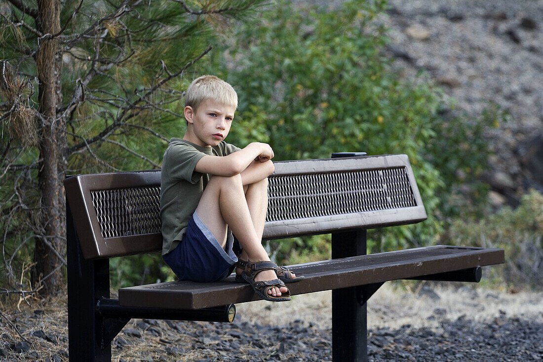 A caucasian boy, 5 to 10, seated on a bench looking unhappy