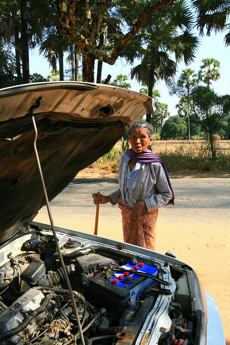 Alte Frau steht hinter offener Kühlerhaube auf einer Landstrasse, Myanmar, Birma, Asien