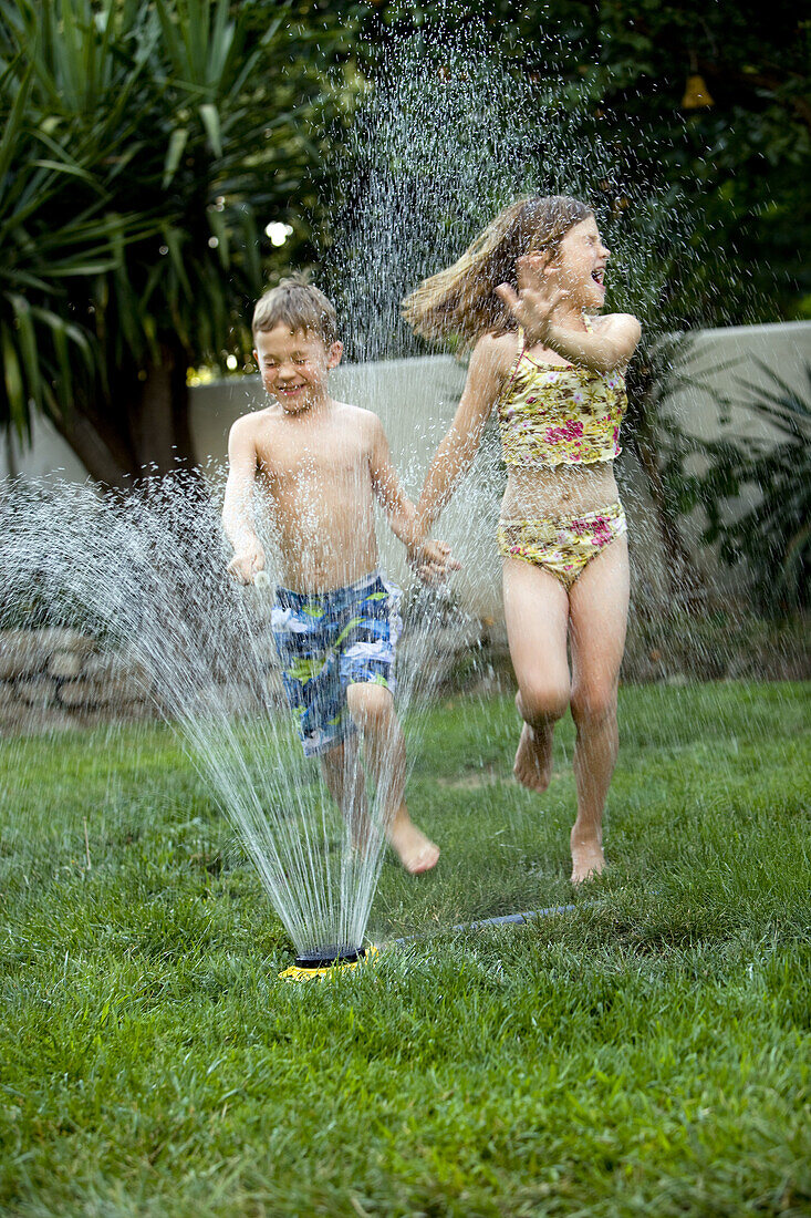 Young boy and girl running through a yard sprinkler