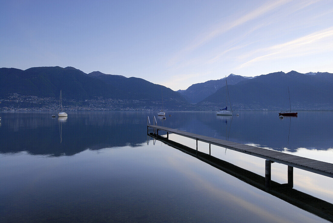 Holzsteg führt in Lago Maggiore mit ankernden Segelbooten mit Locarno, Muralto und Minusio am jenseitigen Ufer, Gambarogno, Lago Maggiore, Tessin, Schweiz