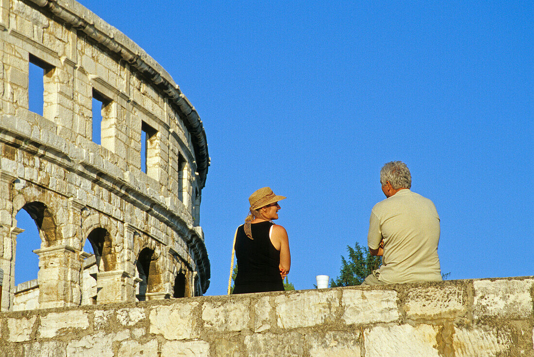 Paar auf der Stadtmauer vor dem römischen Amphitheater unter blauem Himmel, Pula, Kroatische Adriaküste, Istrien, Kroatien, Europa