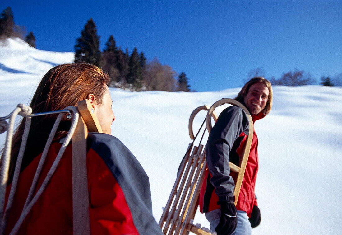 Ein lachendes Paar trägt Schlitten durch den Schnee, Bayern, Deutschland, Europa