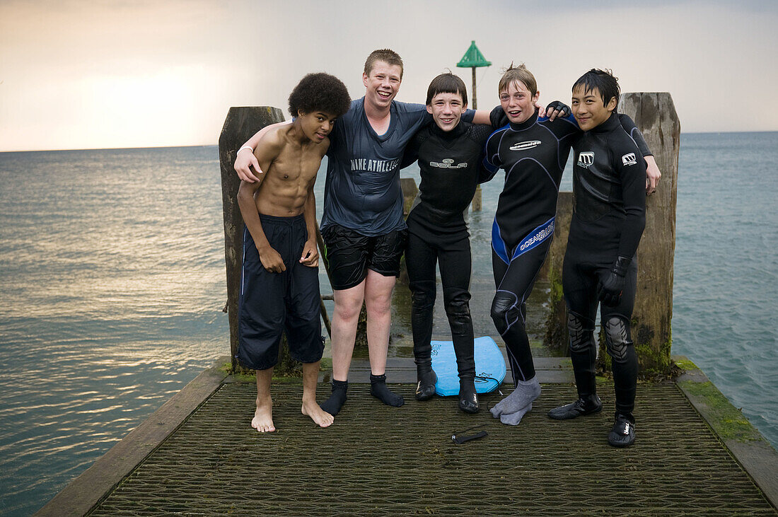 Inter_racial group of five teenage boys, black chinese and white, standing on jetty pier, summer evening, wearing wet suits, Aberystwyth Wales UK