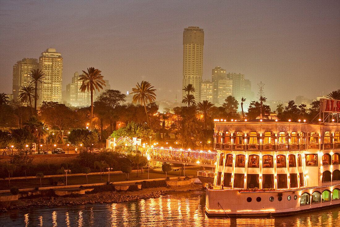 Beleuchtetes Schiff auf dem Nil vor Palmen und Hochhäusern, Kairo, Ägypten, Afrika