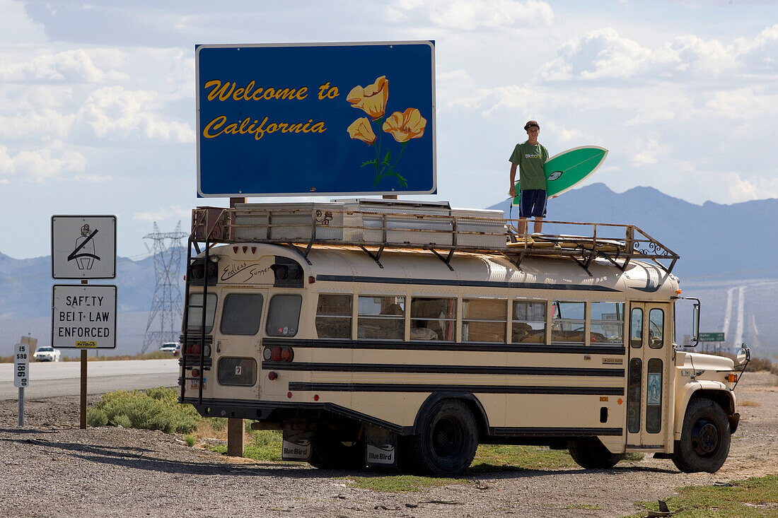 An dem Begrüßungsschild von Kalifornien steht ein 18 Jähriger Junge mit einem Surfbrett auf einem Amerikanischen Schulbus, Interstate 15, Nevada, Kalifornien, USA