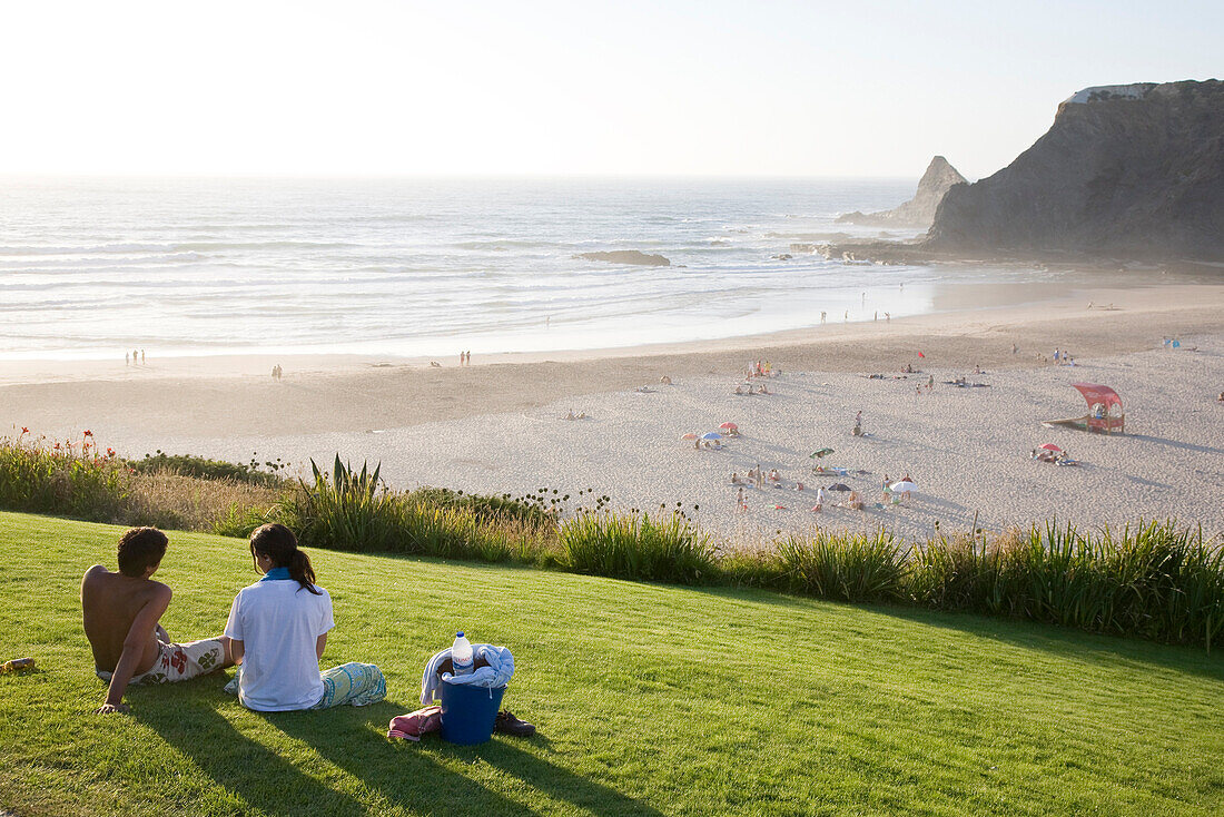 Couple sharing a picnic on a green grass in the evening light, Atlantic ocean, Praia de Odeiceixe, Algarve, Portugal