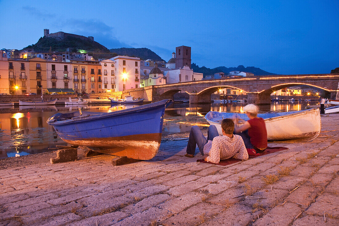 Zwei Touristen am Ufer des Flusses Fiume Temo in der Abenddämmerung, Blick auf das Castello di Serravalle, Bosa, Sardinien, Italien, Europa