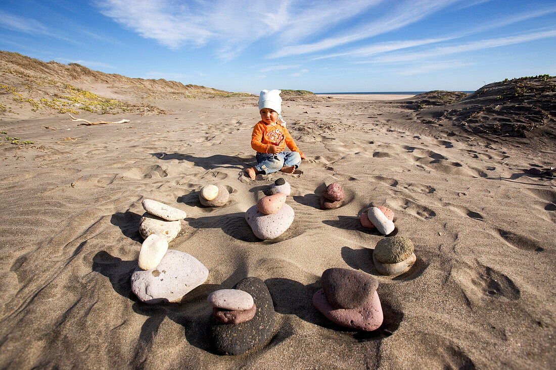 Kleines Mädchen sitzt am Strand vor einem Kreis aus Steinen, Punta Conejo, Baja California Sur, Mexiko, Amerika