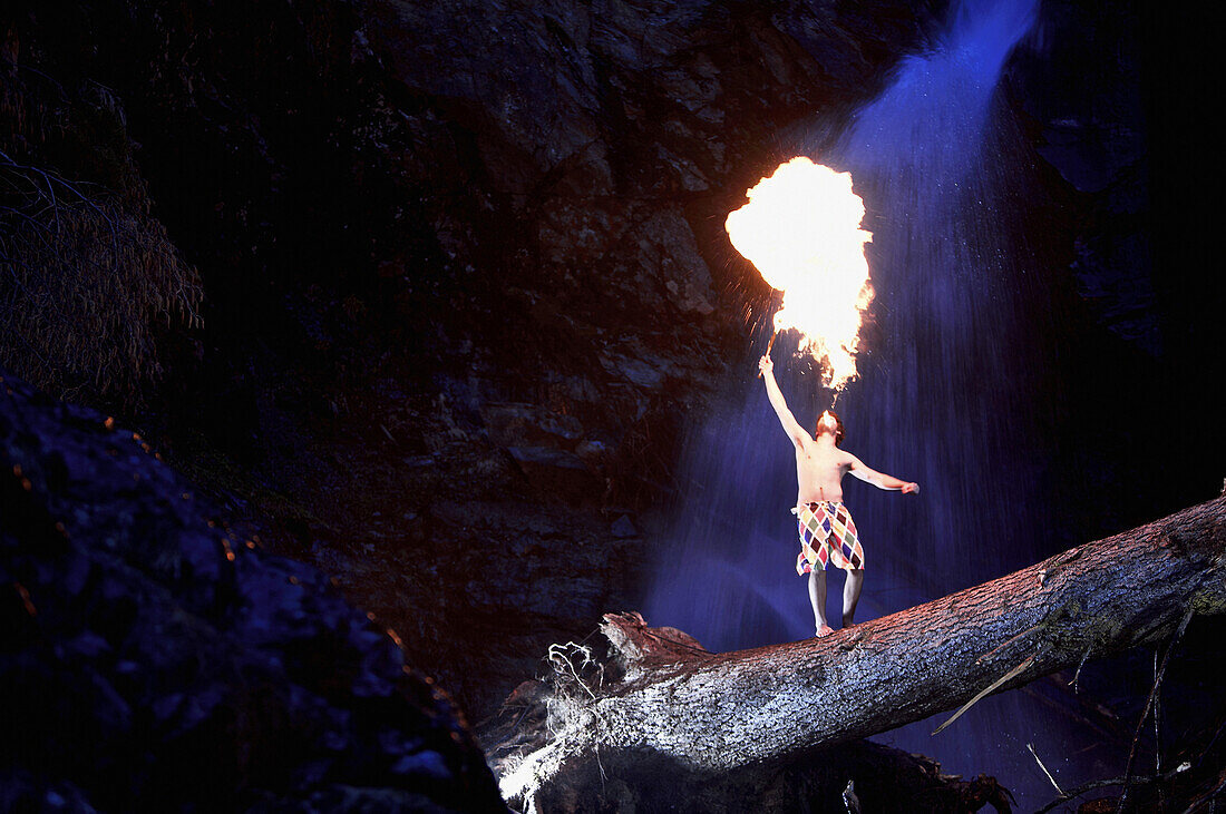 Young man standing on trunk in front of a waterfall while breathing fire, See, Tyrol, Austria