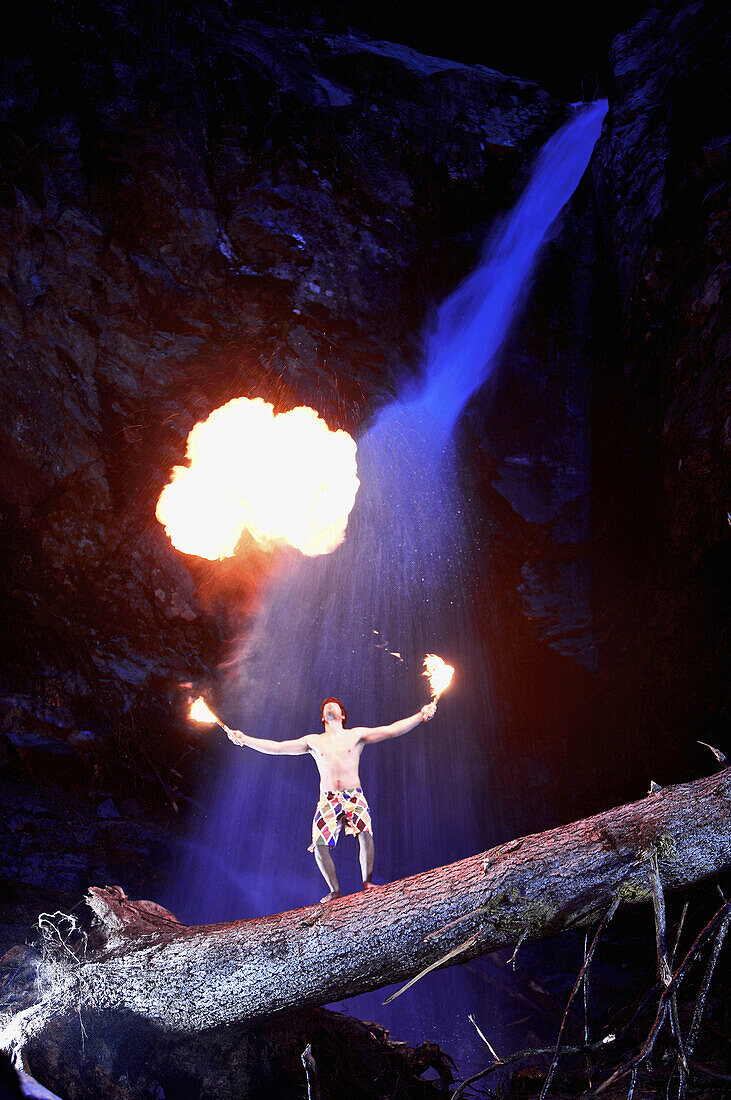 Young man standing on trunk in front of a waterfall while breathing fire, See, Tyrol, Austria