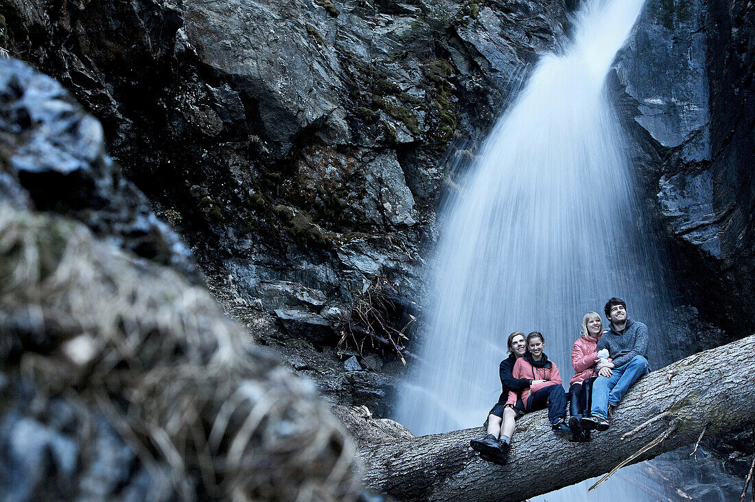 Two couples sitting on a trunk in front of a waterfall, See, Tyrol, Austria