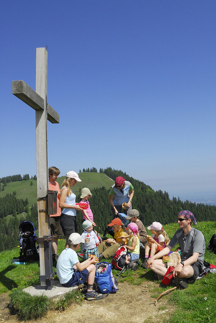 Wandergruppe mit Kindern rastet an einem Gipfelkreuz, Bayerische Alpen, Oberbayern, Bayern, Deutschland