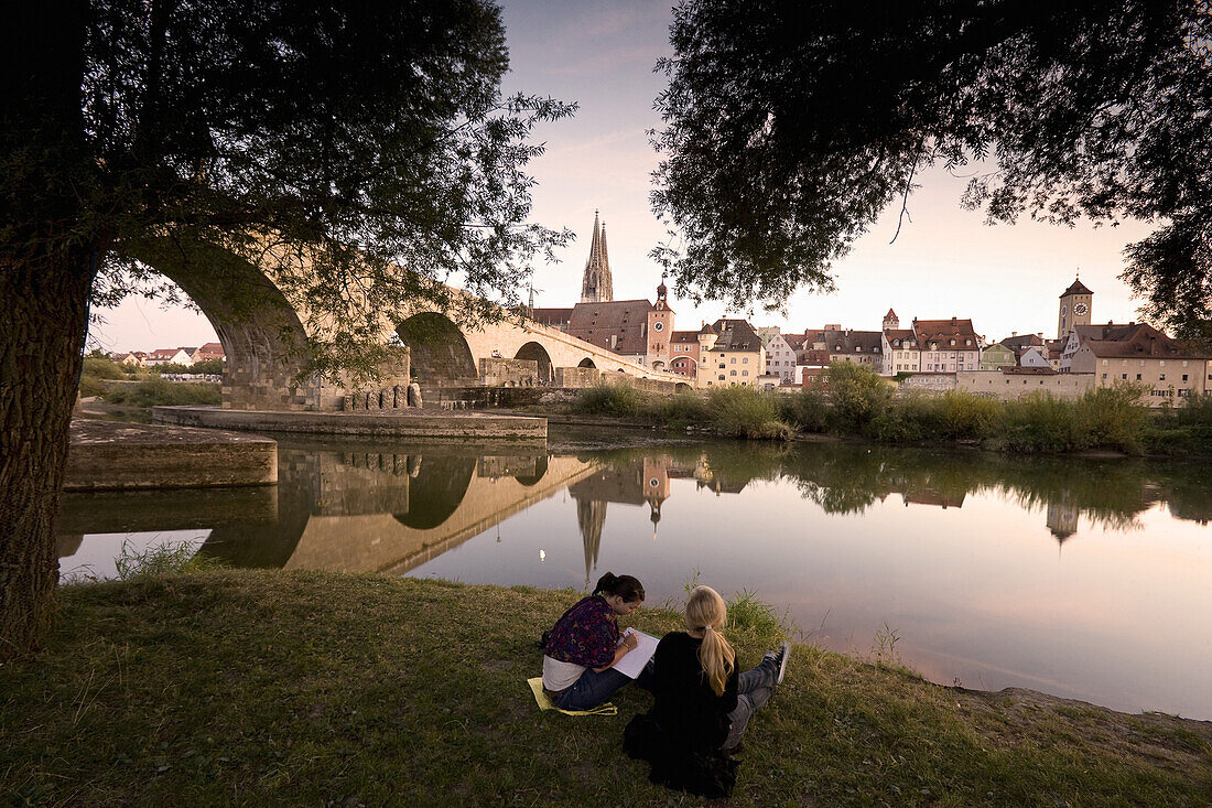 Two young girls sitting on the banks of the river, Stone bridge and Regensburg cathedral, cathedral of St. Peter, Unesco World Cultural Heritage, Donau, Regensburg, Upper Palatinate, Bavaria, Germany, Europe