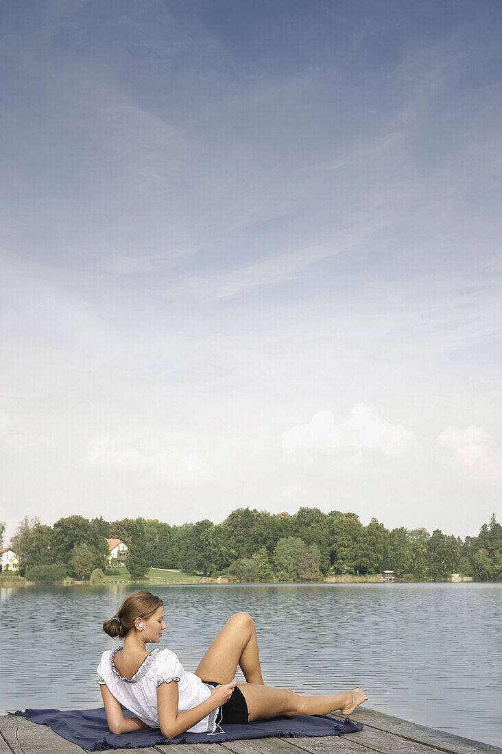 Young woman listening to music while lying on a jetty at lake Starnberg, Bavaria, Germany