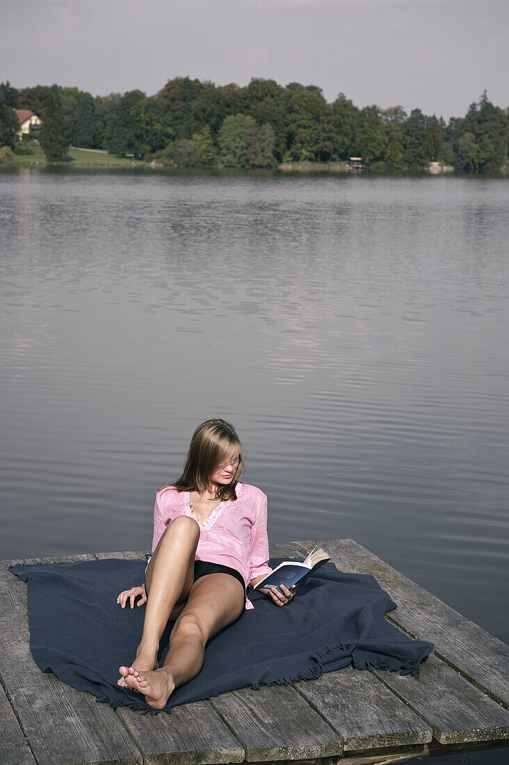 Junge Frau liegt auf einem Steg und liest ein Buch, Starnberger See, Bayern, Deutschland