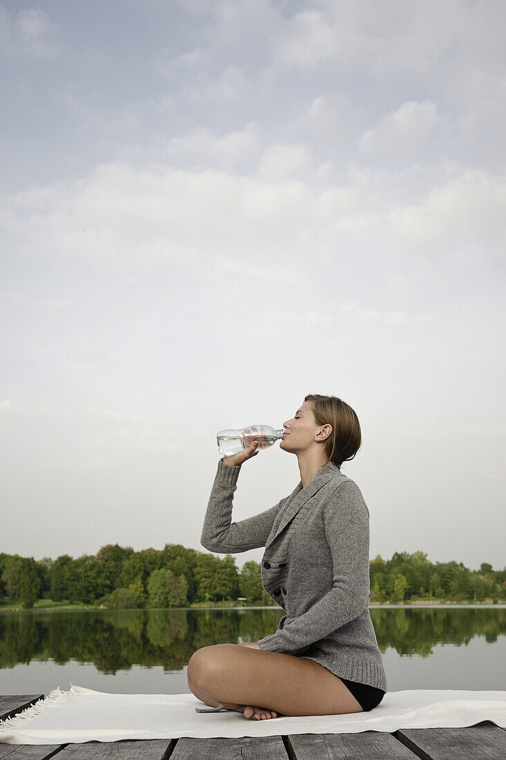 Young woman drinking a bottle of water while sitting on a jetty at lake Starnberg, Bavaria, Germany