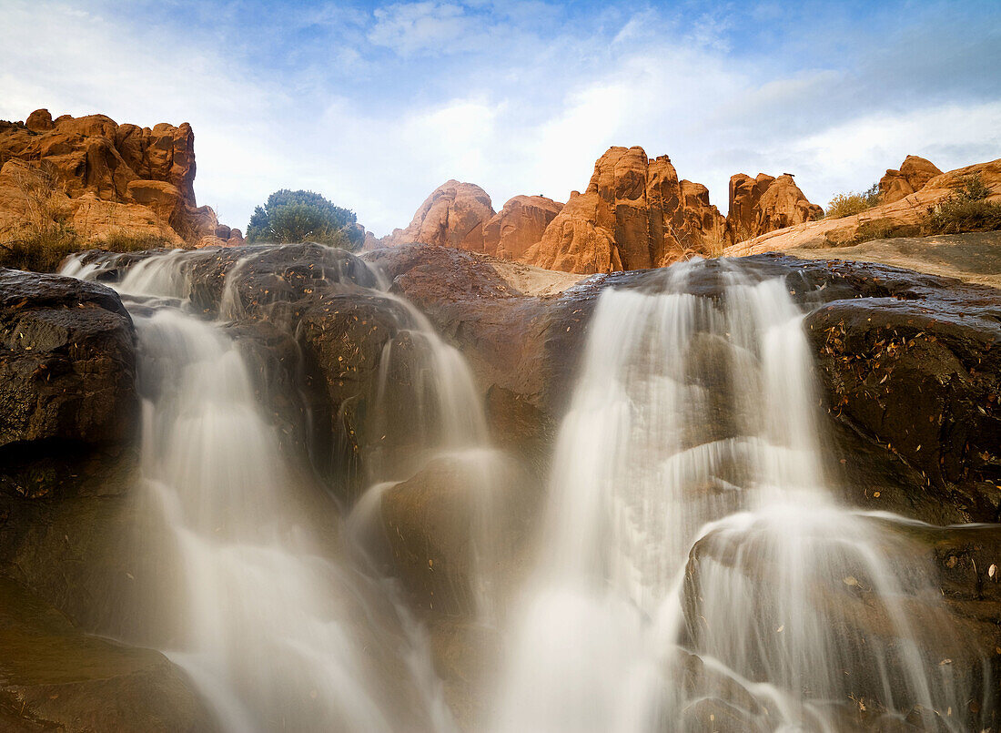 A waterfall cascades below rugged sandstone peaks and a cloud filled sky near Moab,  Utah