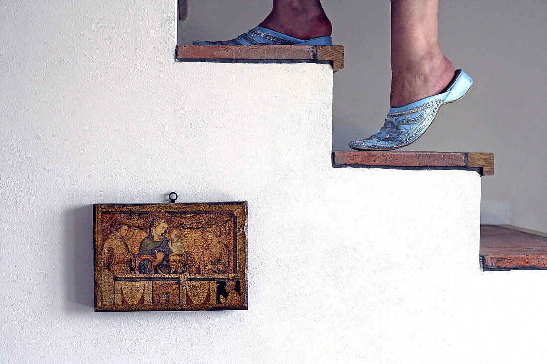 Female feet of a woman wearing traditional maroccan Babush and climbing up the stairs with a picture of a bible scene hanging on the white washed wall of an old landhouse villa,  Gabiano,  Piemont,  Italy,  Europe