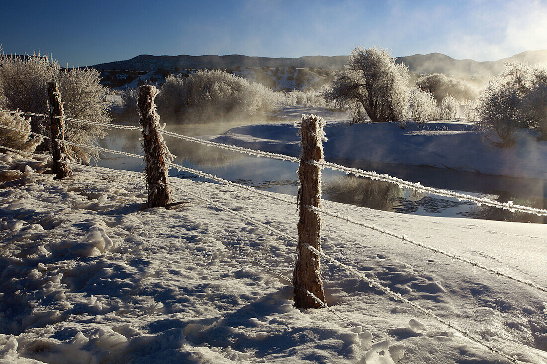 barb wire fence in winter landscape with hoarfrost,  steeming river,  brilliant sunshine and blue sky,  Utah,  USA
