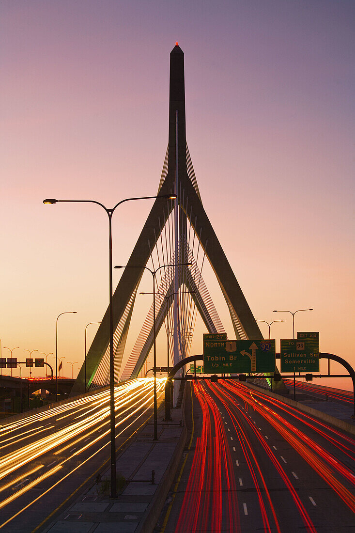 Rush hour traffic light-trails stream in and out of the city of Boston over the Leonard P Kakim Bunker Hill Bridge,  with a colorful sky in the background. Massachusetts,  USA