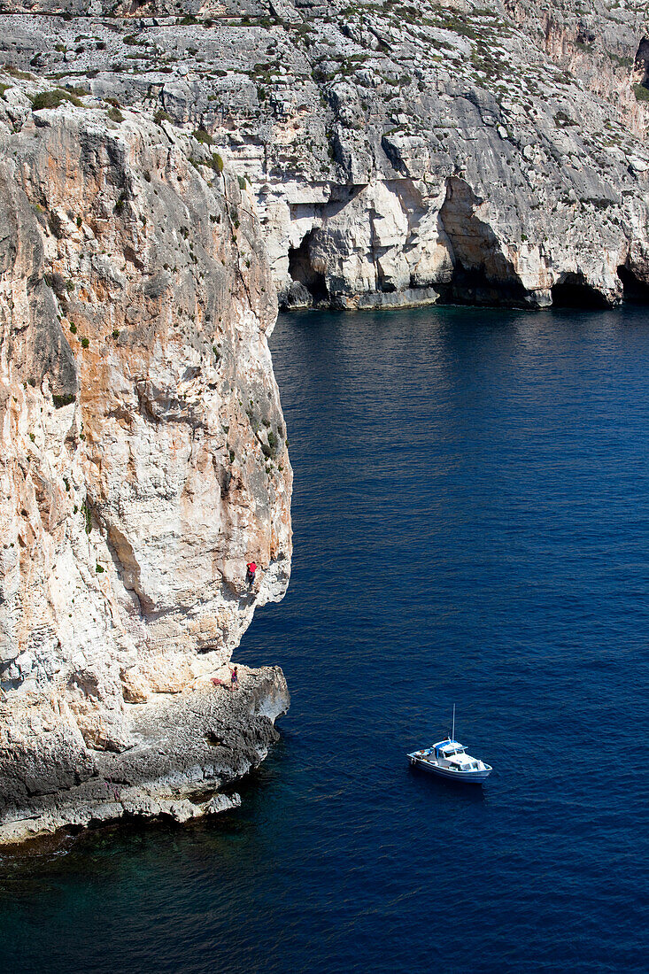 A young woman and a young man climbing on the cliffs at the bay of Zurrieq, a little boat closeby, Malta, Europe