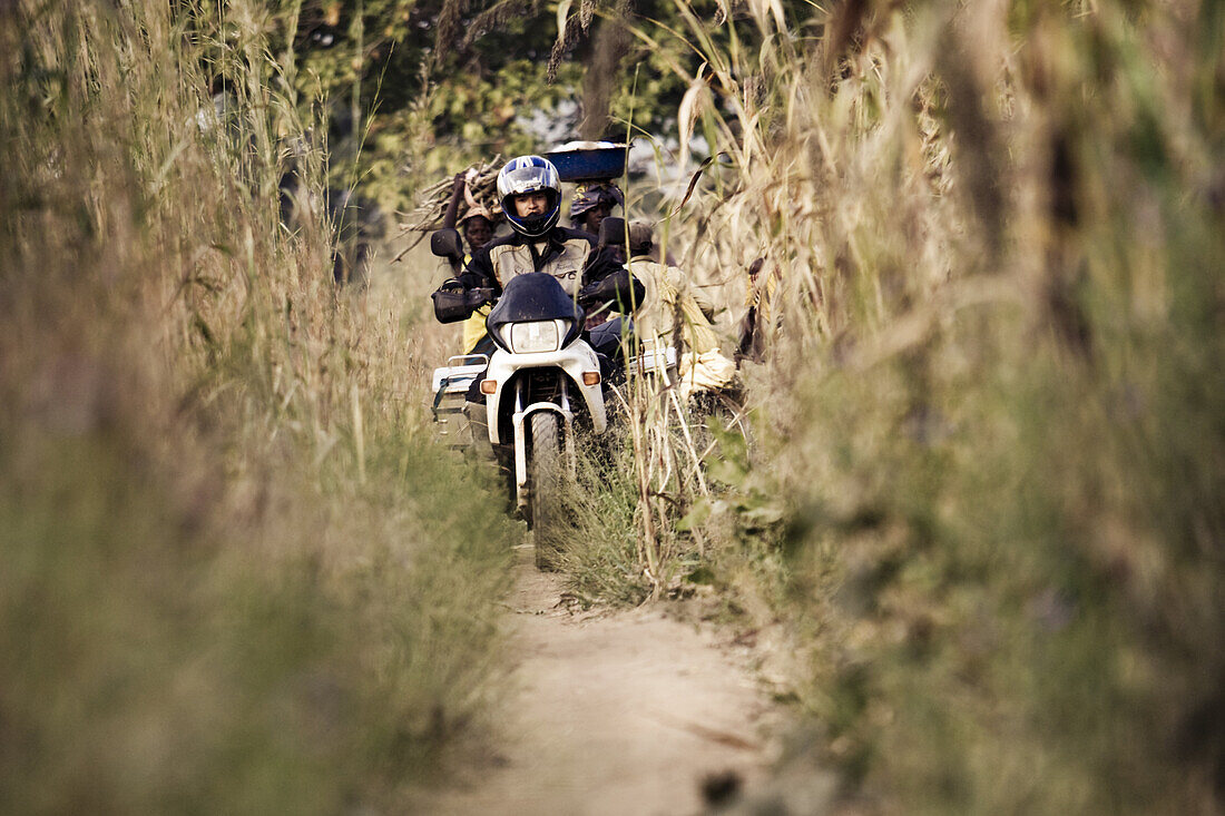 Man on motorcycle driving on narrow path, african women in the background, Siby, Mali, Africa