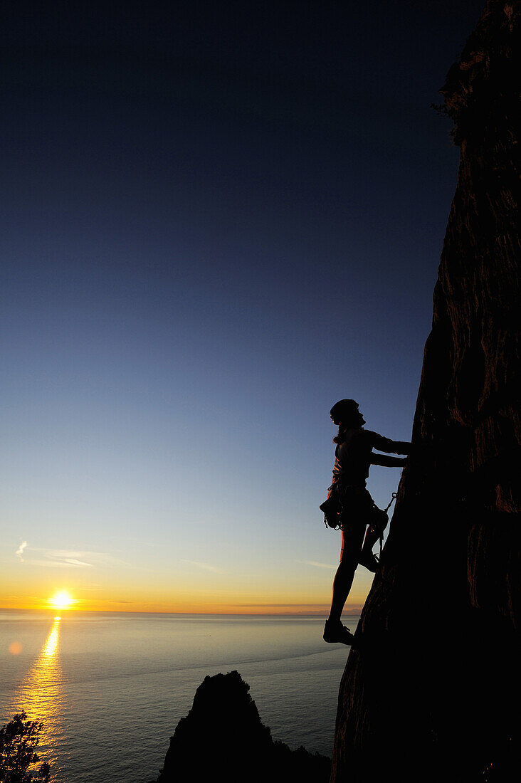 Silhouette einer Frau, Frau klettert an senkrechter Felswand direkt an der Mittelmeerküste, Sonnenuntergang im Meer im Hintergrund, Naturpark Porto Venere, Nationalpark Cinque Terre, UNESCO Welterbe, Ligurien, Italien