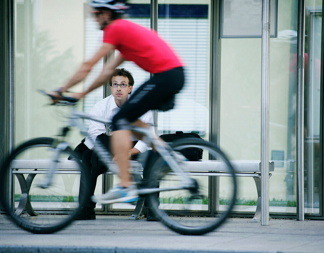 Geschäftsmann sitzt auf einer Bank, Fahrradfahrer fährt vorbei