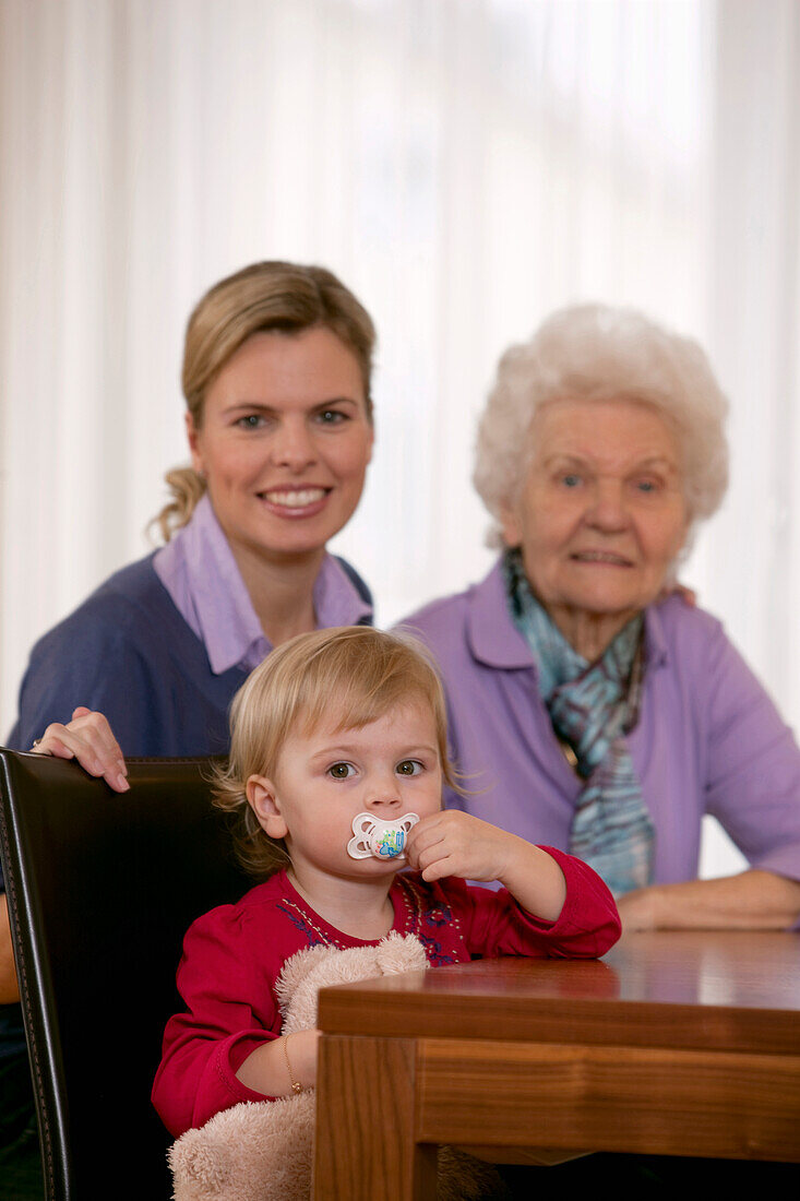 Mädchen (2 Jahre) sitzt mit Mutter und Großmutter an einem Tisch