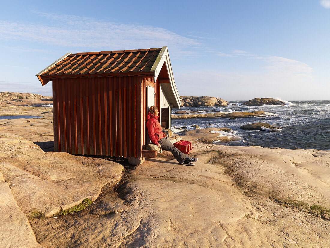 Woman sits by red hut on cliffs, Bohuslan, Sweden
