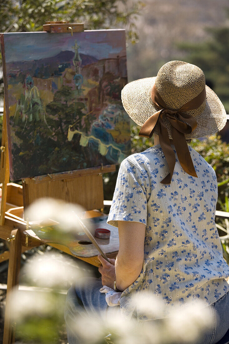 One woman with straw hat and colorful blouse creating landscape oil painting in summer